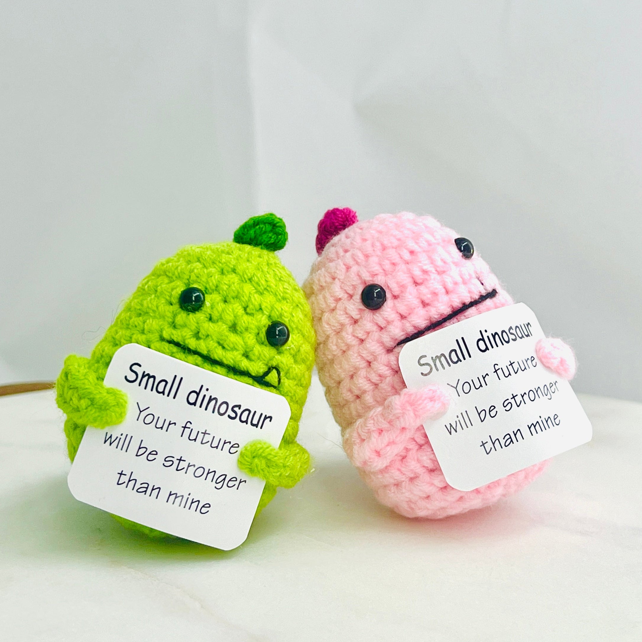Two small crocheted dinosaurs, one green and one pink, with embroidered messages on a white surface.